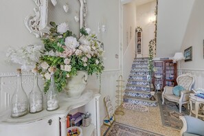 Ein Zimmer mit einem weißen, verzierten Spiegel, einer Blumenkomposition, einer mit blauen und weißen Fliesen ausgelegten Treppe und einem Stuhl mit Kissen.