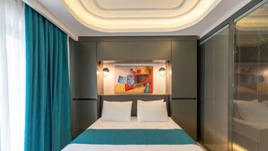A modern hotel room with a large bed, a headboard with artwork, a window with blinds, and a teal curtain. - Gumus Yazar Hotel (Kaş)