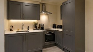 A modern kitchen with dark cabinets, a stainless steel range hood, a built-in oven, and a countertop with various appliances. - Opulenti Mayfair 1BR Residence (London)