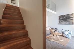 Un escalier en bois mène à un espace de vie comprenant un canapé, un fauteuil à bascule et une œuvre d’art accrochée au mur.