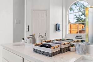 A bathroom with a white countertop, a mirror, a towel rack, and a window with a view of a wooden deck. - Modern Oasis with Large Pool + Ping-Pong Sleeps 14 (McKinney)