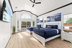 A bedroom with two beds, a ceiling fan, a large window, and a painting on the wall. - Modern Oasis with Large Pool + Ping-Pong Sleeps 14 (McKinney)