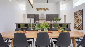 A modern dining area with a wooden table, black chairs, and a kitchen in the background. - Avantstay Polo Villa Halo Pickleball, Putting Green Sleeps 28 (La Quinta)