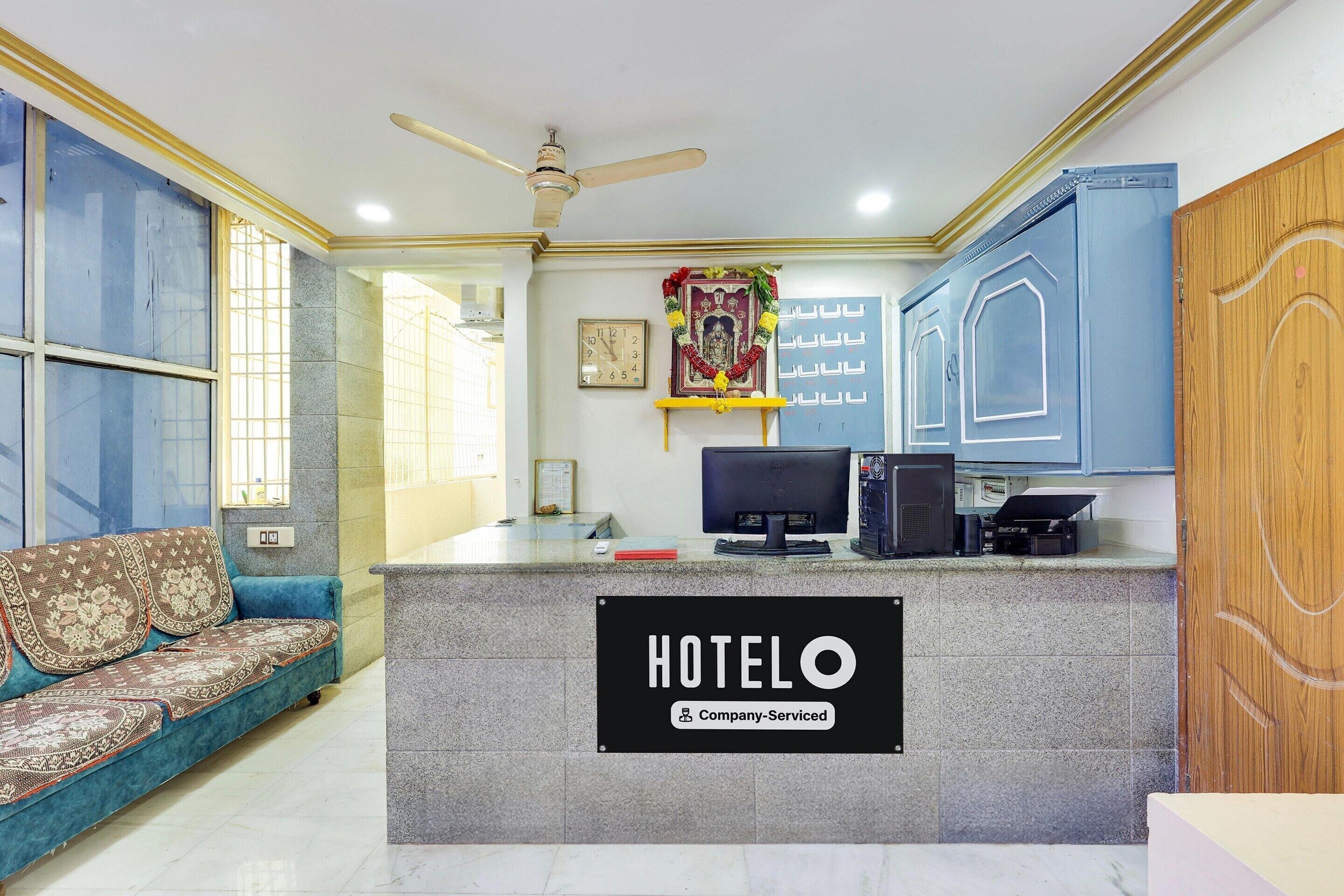 A reception desk with a sign reading 'HOTELO Company Serviced,' a ceiling fan, a clock, and a blue sofa.