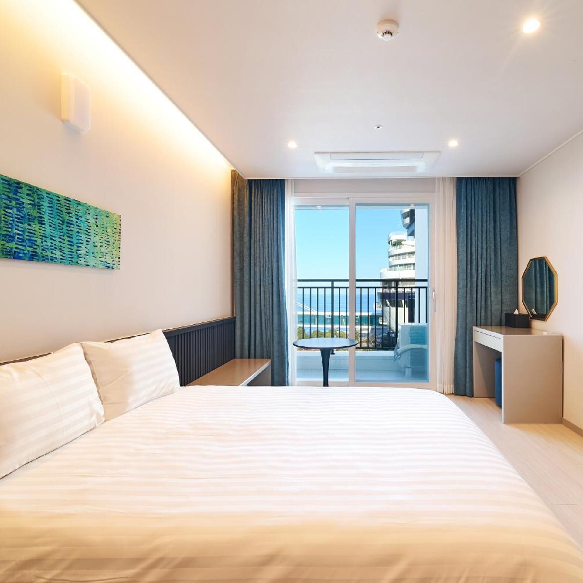 A hotel room with a bed, a small table, a desk, a chair, a balcony with a railing, and a view of a building.