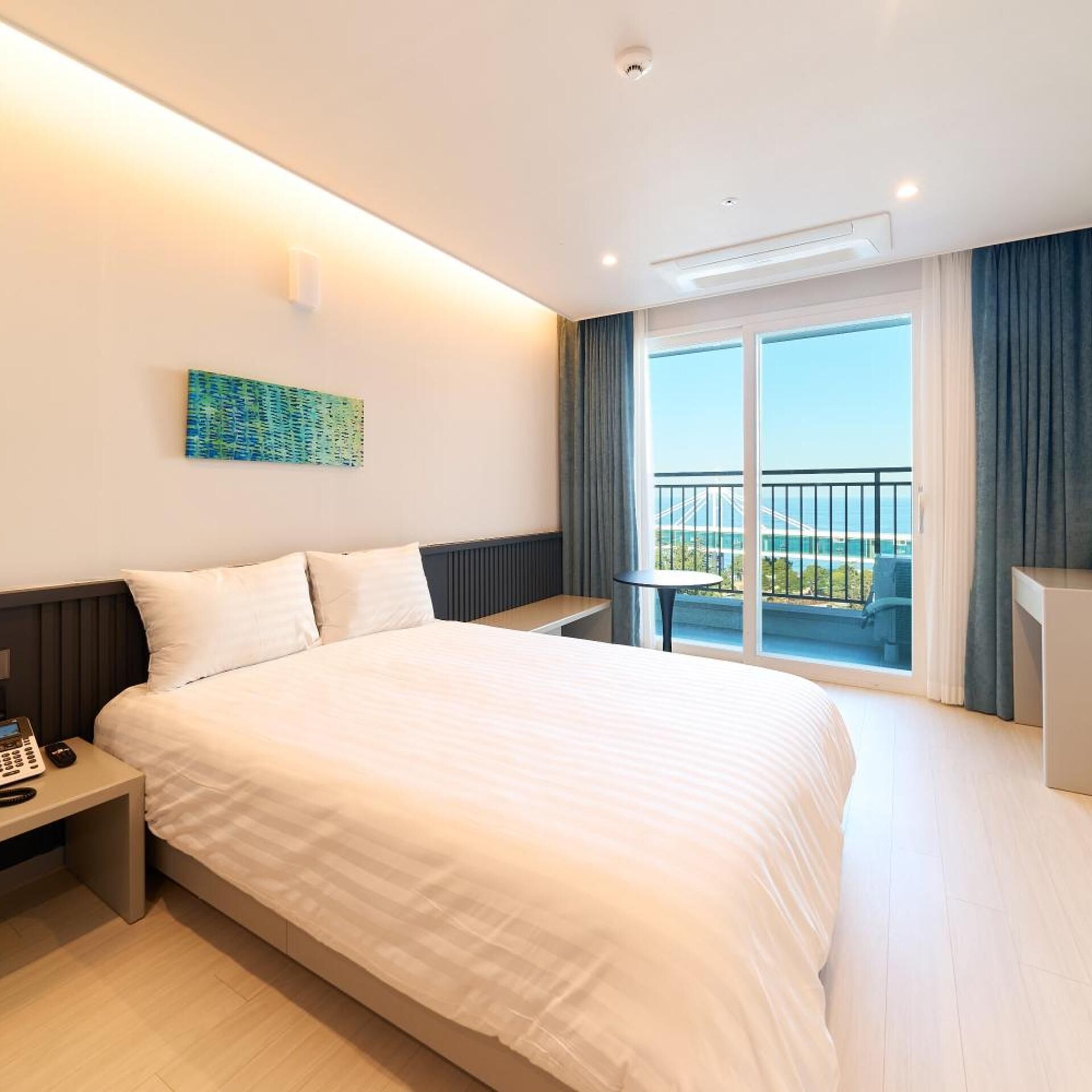 A hotel room with a large bed, a bedside table with a phone, a small table with a chair, and a balcony with a view of the sea and a bridge.