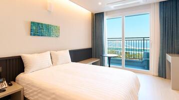 A hotel room with a large bed, a bedside table with a phone, a small table with a chair, and a balcony with a view of the sea and a bridge.