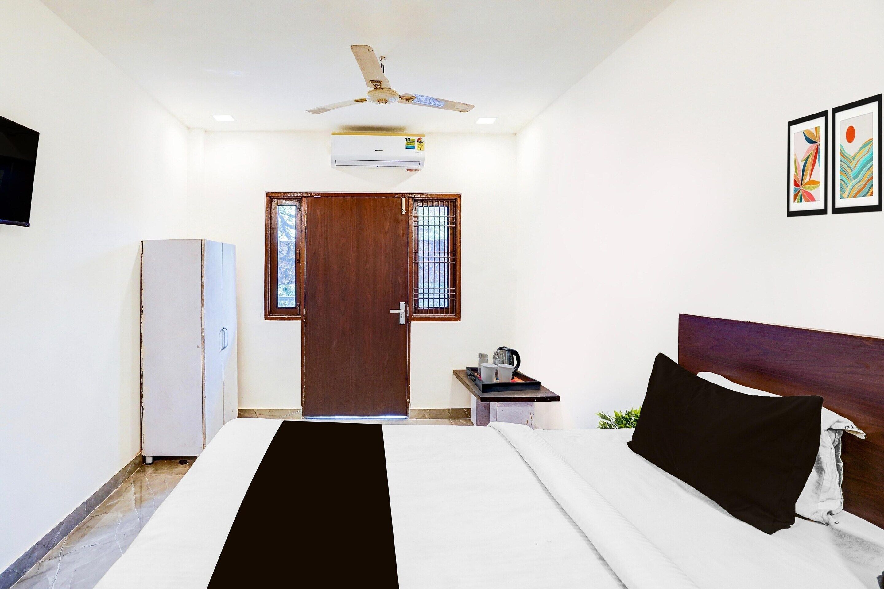 Une chambre d’hôtel avec un lit, un ventilateur de plafond, un climatiseur, une porte, une petite table avec une bouilloire et un téléviseur fixé au mur.