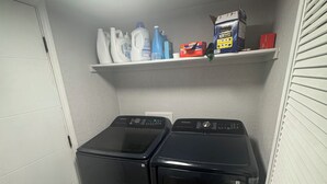 Een wasserette met twee voorladers wasmachines en drogers, een plank met schoonmaakmiddelen, en een witte deur.
