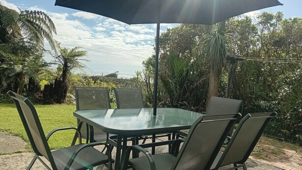 Eine Terrassengruppe im Freien mit Glastisch und Stühlen, einem großen Sonnenschirm und üppigem Grün.