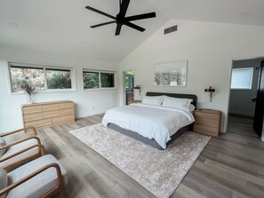 Makuuhuone, jossa on sänky, yöpöydät, kattotuuletin, ikkunat, lipasto ja matto.