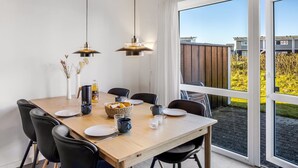 En matplats med ett träbord, svarta stolar, hängande taklampor och utsikt över en trädgård genom stora fönster.