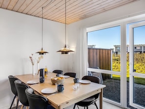 A dining area with a wooden table, black chairs, hanging pendant lights, and a view of a garden through large windows.