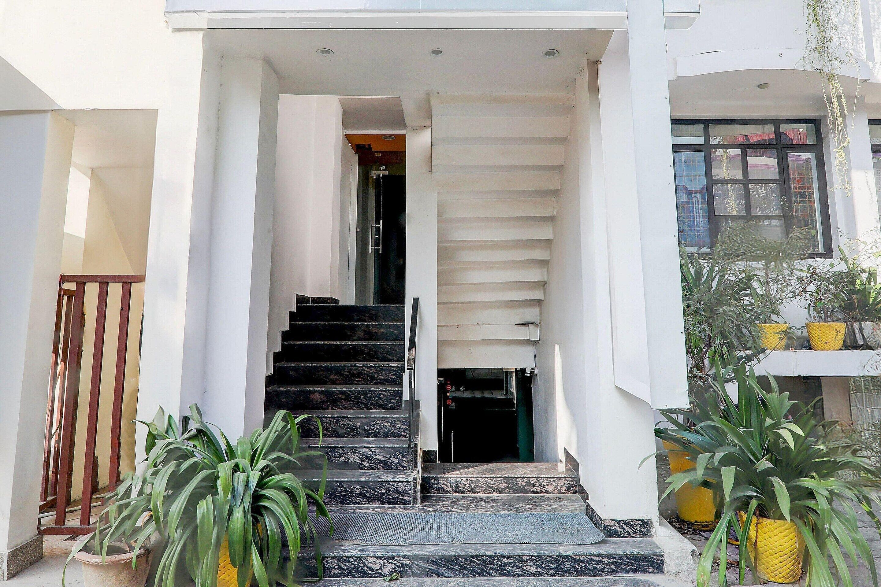 Entrada de edifício com escadas que levam a uma porta, plantas em vasos e corrimão de metal.