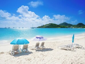 A beach with white lounge chairs and umbrellas, clear turquoise water, and a distant green hill.