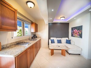 Eine moderne Küche mit Holzausstattung, einem Gaskochfeld und einer Arbeitsfläche. Im Wohnbereich befindet sich ein weißes Sofa mit blauen Kissen und ein Couchtisch. An der Wand hängt ein Gemälde von Korallen.