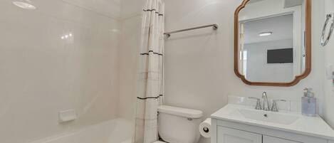 A bathroom with a white toilet, a shower curtain, a bathtub, a sink with a mirror, and a light fixture.
