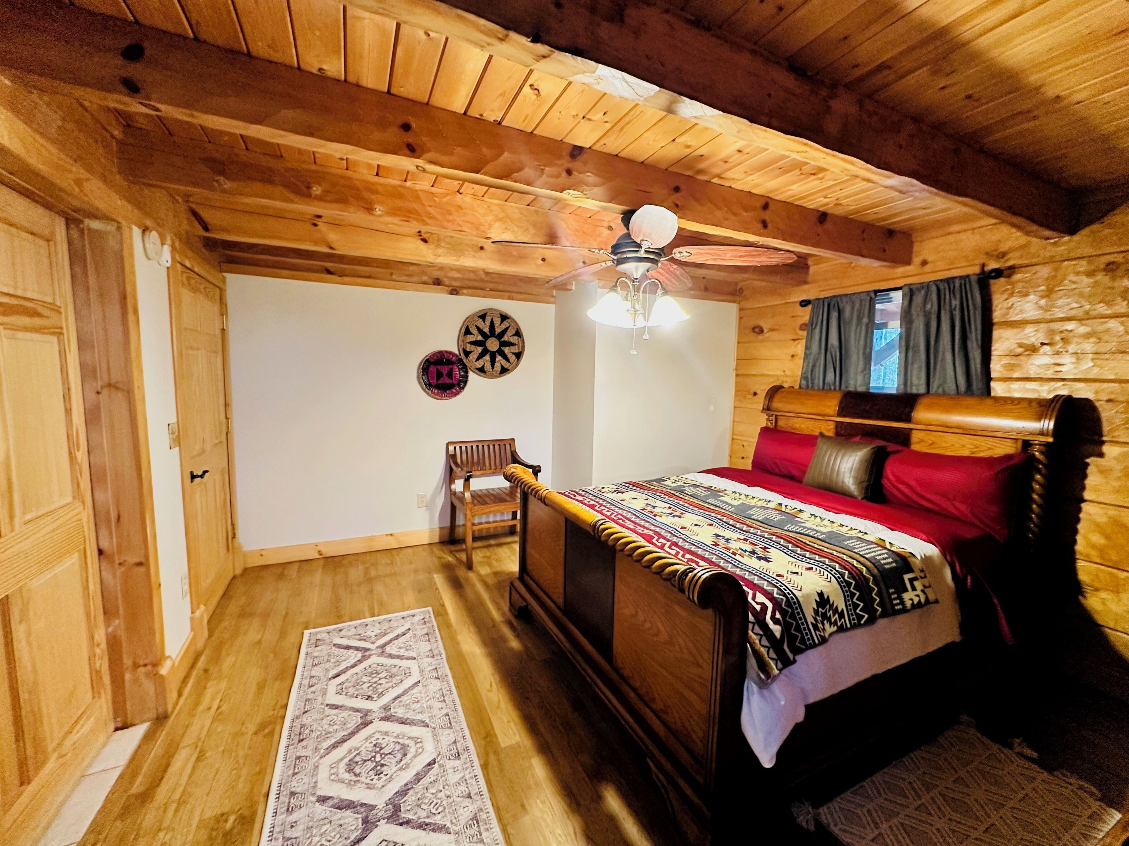 A wooden cabin bedroom with a bed, a chair, a ceiling fan, and a wall-mounted lamp.