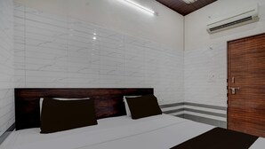 A hotel room with a large bed, a wooden headboard, white tiled walls, a wooden door, and an air conditioning unit. - Deluxe Krishna Janam Bhoomi (Mathura)