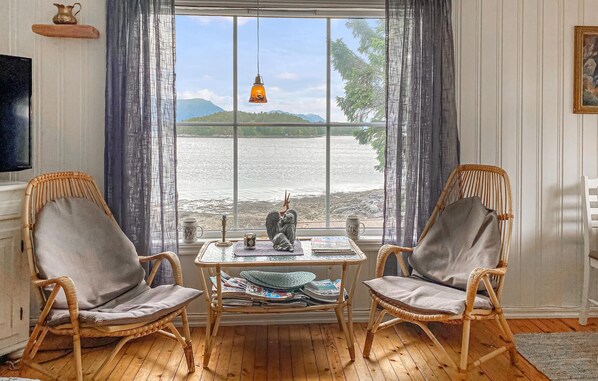 A room with two wicker chairs, a small table with newspapers, a decorative pitcher, and a view of a lake and mountains.