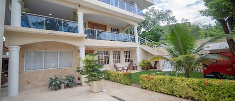 A multi-story building with balconies, a garden area with plants and flowers, and a red vehicle parked outside.