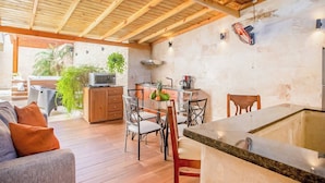 Un grand espace extérieur aménagé en cuisine, avec un sol en bois, une table à manger et des chaises, une cuisinière encastrée et un téléviseur.