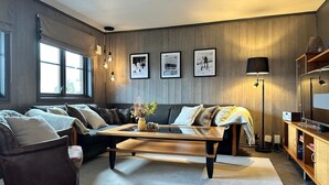 Una sala de estar acogedora con un sofá, sillones, una mesa de centro y una chimenea. Hay cuadros enmarcados en la pared y una lámpara en el suelo.