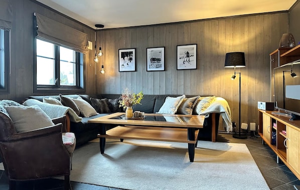 A cozy living room with a sofa, armchairs, a coffee table, and a fireplace. There are framed pictures on the wall and a lamp on the floor.