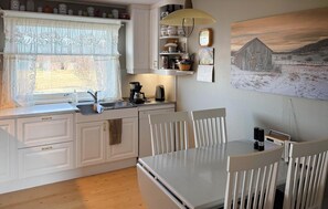 Une cuisine équipée d’un évier, d’une cuisinière et de meubles blancs. On y trouve une table à manger avec des chaises, ainsi qu’un tableau accroché au mur.