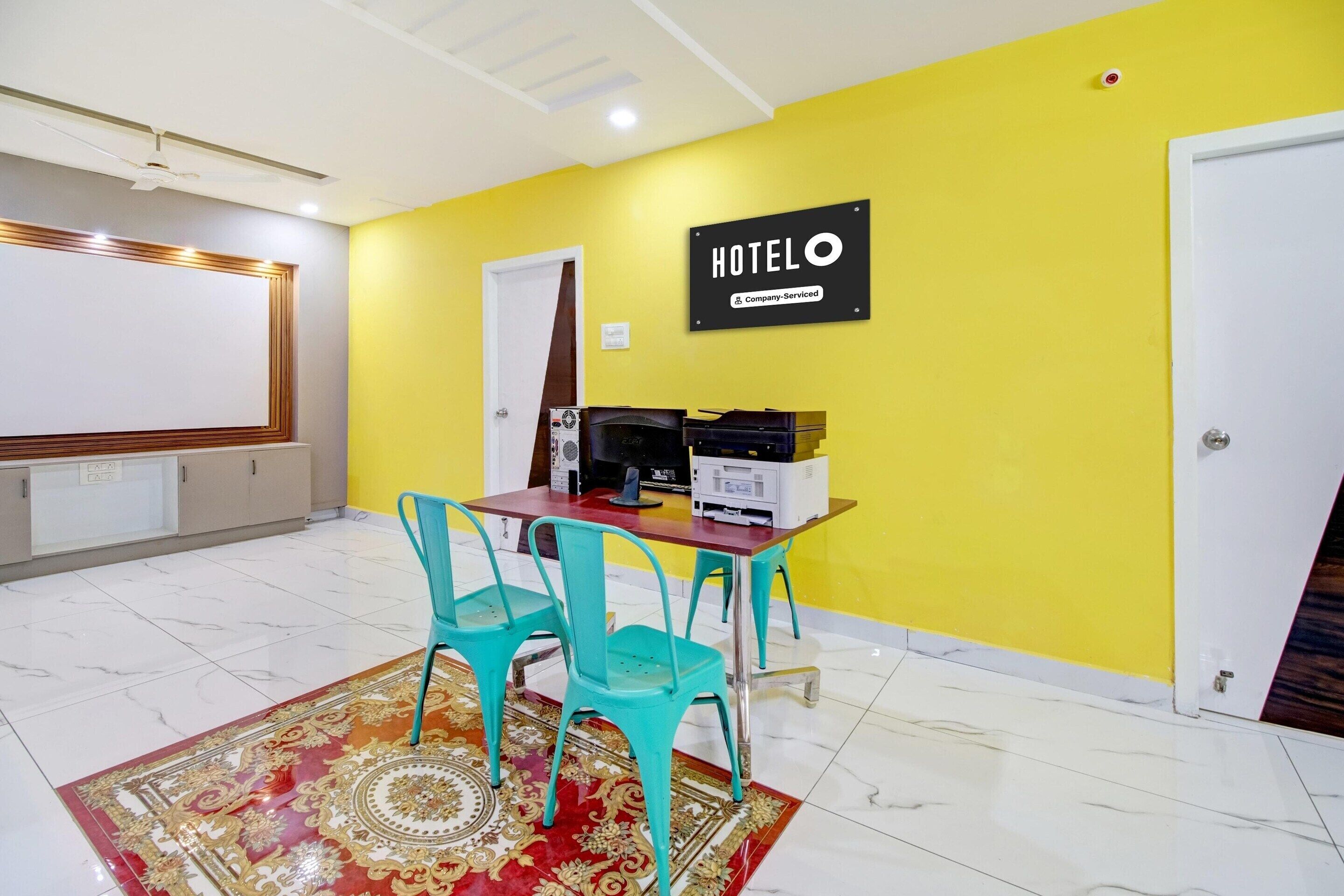 Una habitación de hotel con una pared amarilla, un letrero que dice 'HOTEL', una puerta blanca, una mesa con sillas, una pantalla de proyector y un ventilador de techo.