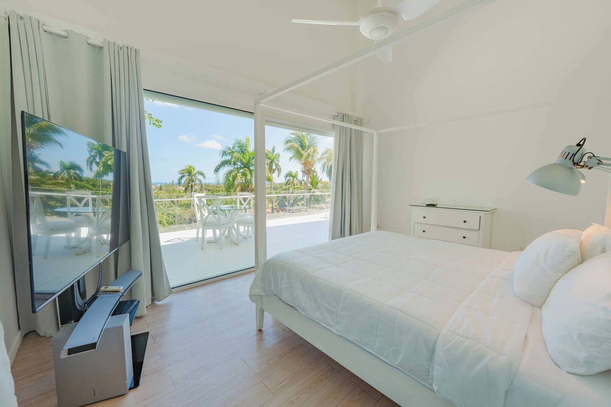 Quarto com cama, escrivaninha, cadeira, TV, ventoinha no teto e varanda com vista para palmeiras.