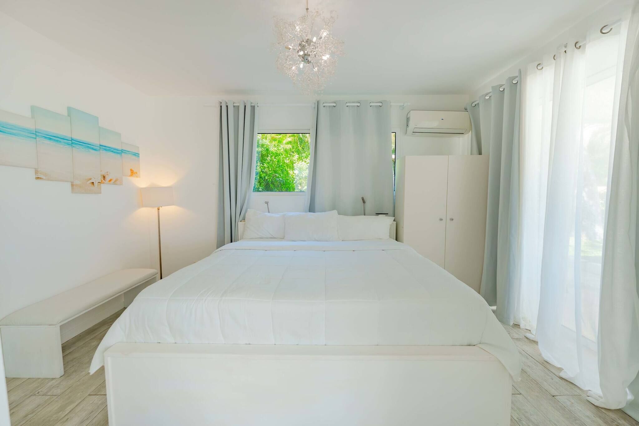Une chambre avec un grand lit, un banc, un climatiseur fixé au mur et une fenêtre avec des rideaux.