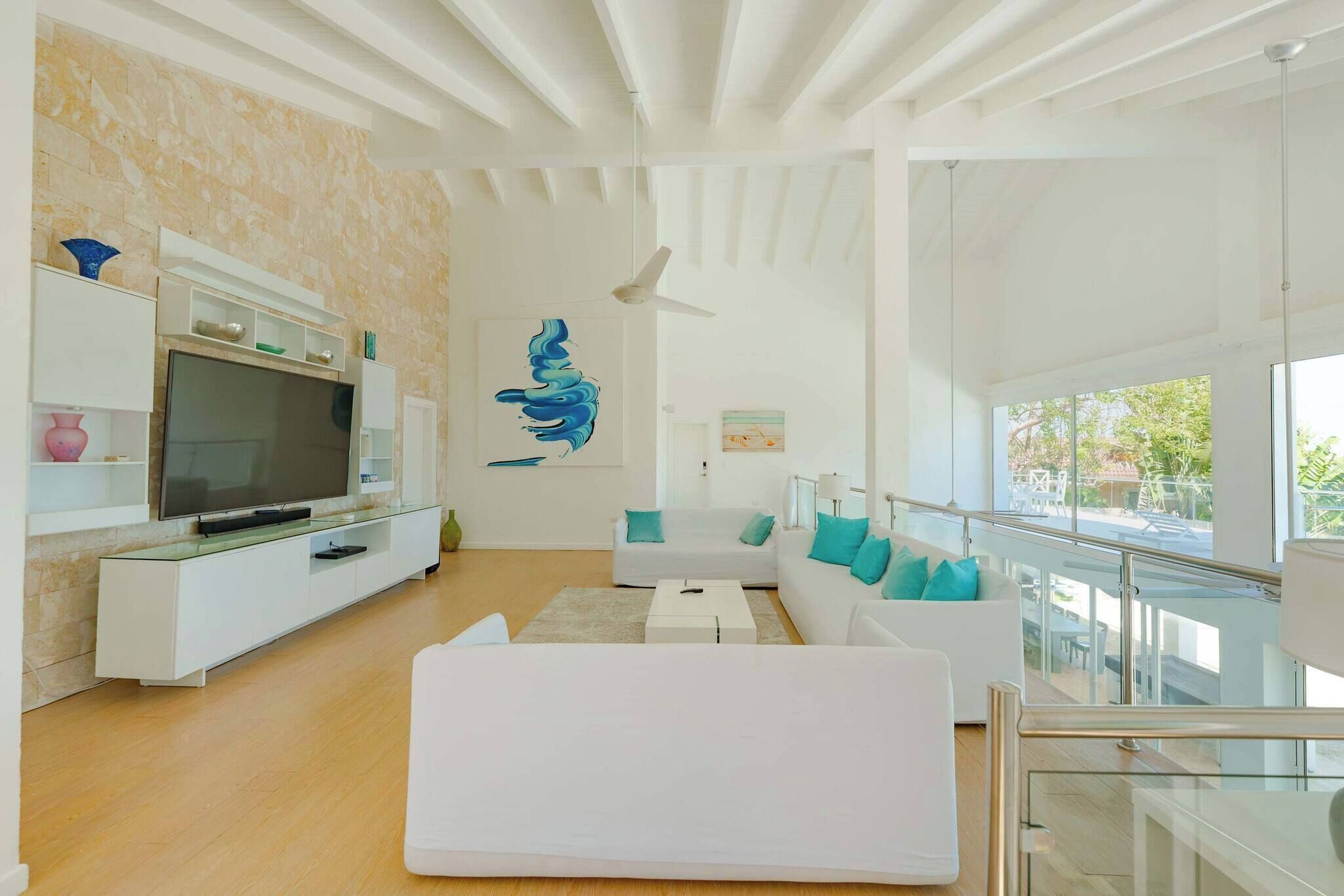 A modern living room with a flat-screen TV, white sofas, a glass railing, and a ceiling fan.