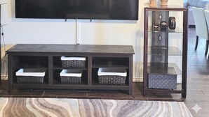 A living room with a flat-screen TV mounted on the wall, a dark wooden entertainment unit with storage baskets, and a glass display cabinet with decorative items. - Spacious 4-bedroom 2 bath house equipped with AC. Sleeps up to 12 people. (Lawton)