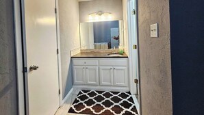 A narrow hallway leading to a bathroom with a white vanity, a mirror, and a patterned rug. - Spacious 4-bedroom 2 bath house equipped with AC. Sleeps up to 12 people. (Lawton)