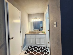 Un couloir étroit mène à une salle de bain équipée d’un meuble-lavabo blanc, d’un miroir et d’un tapis à motifs.