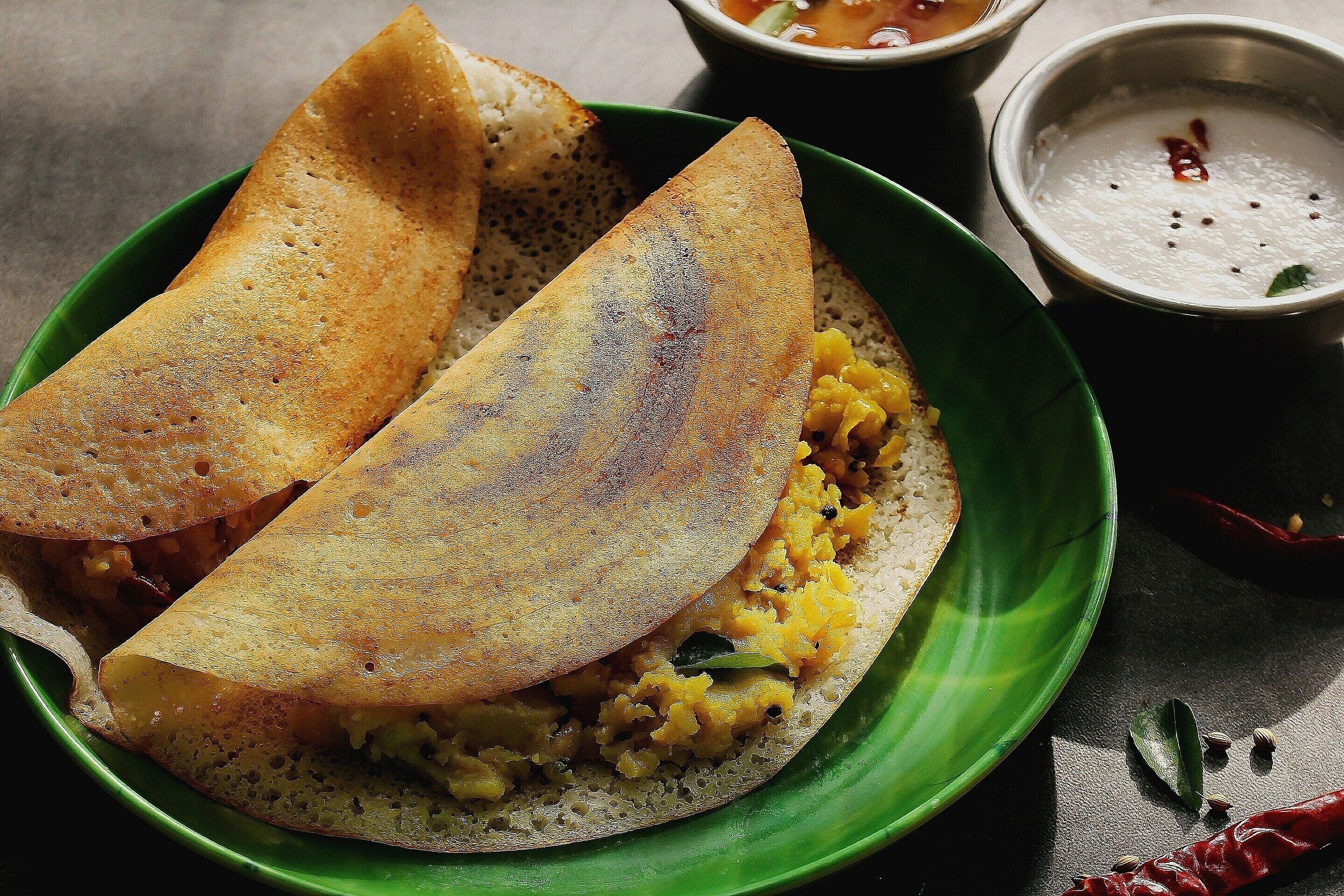 A plate with a folded crepe and a green bowl containing a yellow dish, possibly a curry, with a small bowl of white sauce and a red chili.