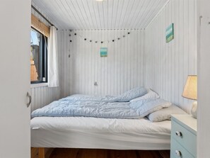 Ein Einzelbett mit weiß-blauer Bettdecke, ein Nachttisch mit Lampe und ein Fenster mit Vorhang.