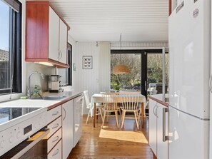 A kitchen with wooden cabinets, a dining table with chairs, a refrigerator, and a window with a view of trees. - "Emelita" - 1.1km from the sea by Interhome (Græsted)
