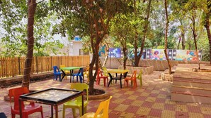 Un espacio al aire libre con mesas y sillas, rodeado de árboles y un muro decorativo.