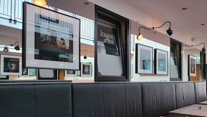 Een restaurantinterieur met zwarte lederen zithoeken, houten tafels en ingelijste kunstwerken aan de muren.