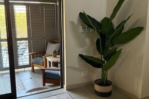 Ein Zimmer mit einer Schiebetür, die auf einen Balkon führt, einer Kübelpflanze, einem kleinen Tisch mit Lampe und einem Sessel mit Kissen.