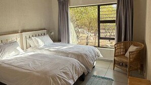 A bedroom with two beds, a window with curtains, a wicker chair, and a view of trees. - Ingonyama Rest (Marloth Park)