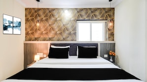 A hotel room with a bed, a window, a wall with a geometric pattern, and a nightstand with a lamp. - Deluxe Hotel Blue Petals (Hyderabad)