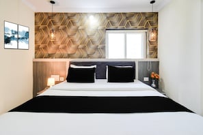 Una habitación de hotel con cama, ventana, pared con diseño geométrico y mesita de noche con lámpara.