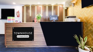 A reception desk with a sign reading 'TOWNHOUSE Company Serviced,' a flat-screen TV, a fire extinguisher, and a potted plant. - Deluxe Hotel Blue Petals (Hyderabad)