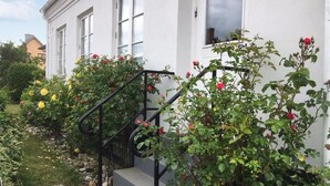 En vit byggnad med en svart trappa som leder till en dörr, omgiven av grönska och blommor.