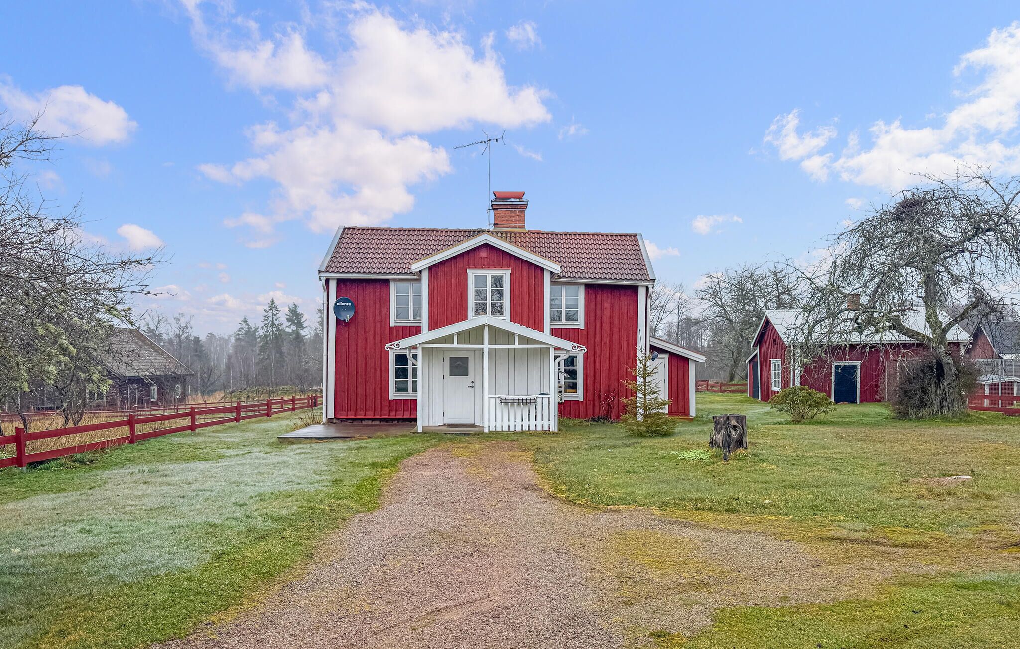 A red farmhouse with a white porch, a gravel driveway, and a fenced yard.
