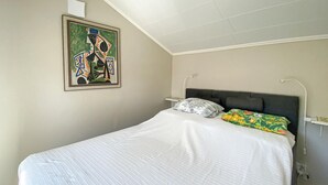 Un dormitorio con cama, cabecera, mesita de noche, lámpara de pared y un cuadro abstracto enmarcado.