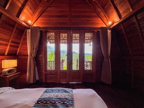A room with a bed, wooden walls, a balcony with curtains, and a view of greenery.